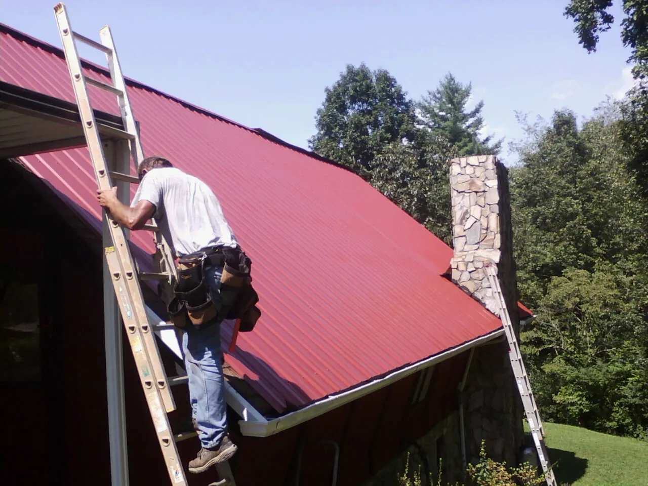 Roofer on a ladder beside a red metal roof and stone chimney — Soffit & Fascia Repair by Roofing Brothers in Herman